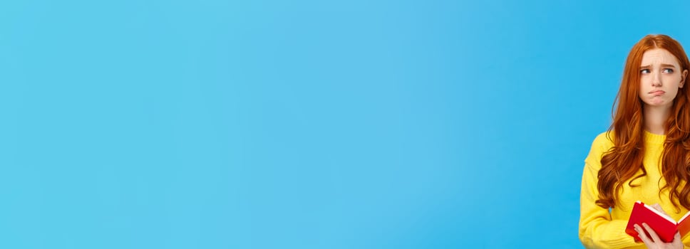 Troubled and pensive uneasy pretty redhead female, pouting and frowning looking away thoughtful, feeling sad and distressed, reading something bad in notebook, standing blue background photo