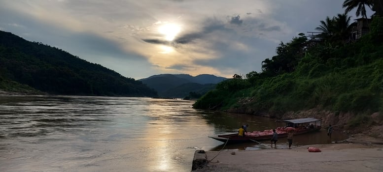 Sunset over a calm river with boats and lush greenery on the shore, surrounded by mountains. photo