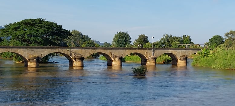 Historic stone bridge with archways spanning a tranquil river in a picturesque rural setting, surrounded by lush greenery and clear blue sky. photo