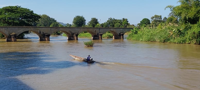 A small boat navigates a serene river under a historic stone bridge, surrounded by lush greenery and a clear blue sky. photo