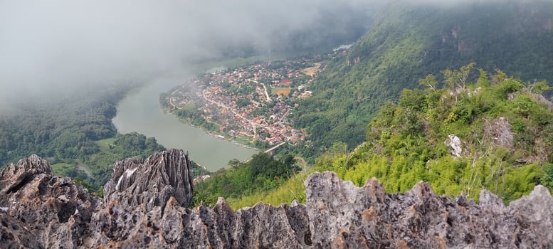 A breathtaking view from a rocky mountaintop overlooking a river and small village nestled among lush, green hills under a cloudy sky. photo