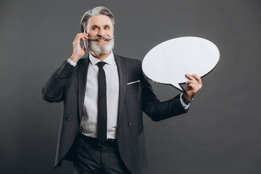 Business and fashionable bearded mature man in a gray suit talking by phone, smiling and holding white paper blank for the copy space on the grey background. photo