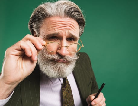 Bearded and mustachioed middle-aged teacher in khaki suit holds hand with glasses and looks closeup on green background photo