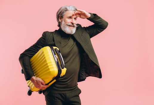 Bearded gray-haired middle-aged man in a suit hold a yellow suitcase on a pink background photo