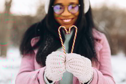 Young african american woman in glasses and warm mittens holding two heart shaped sweet candies on winter background on Valentines Day photo