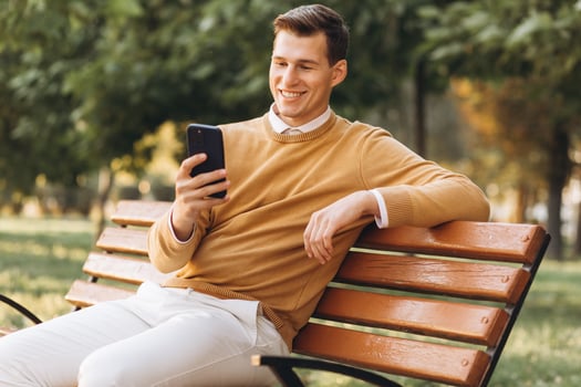 Modern handsome smiling man in yellow and white clothes talking on the phone sitting on a park bench at sunset photo