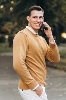 Modern handsome smiling man in yellow and white clothes talking on the phone in the park at sunset photo