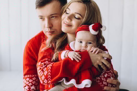 Portrait of happy family mom dad and little daughter in red traditional christmas clothes spending time together in light wooden room near window, copy space photo