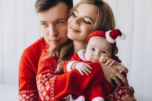 Portrait of happy family mom dad and little daughter in red traditional christmas clothes spending time together in light wooden room near window, copy space photo