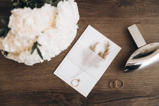 Wedding invitation or greeting card mockup with craft envelope, parfume, white peony flowers bouquet and wedding rings on wooden background. photo