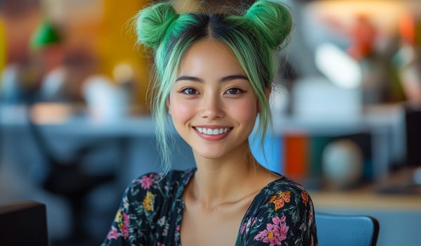 A woman with green hair and a floral dress is smiling. She is sitting at a desk with a laptop photo