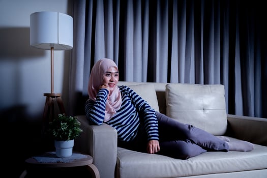 Asian muslim woman wearing hijab sitting using the laptop to do work, working from home at her house on the sofa and sit and watch television. photo