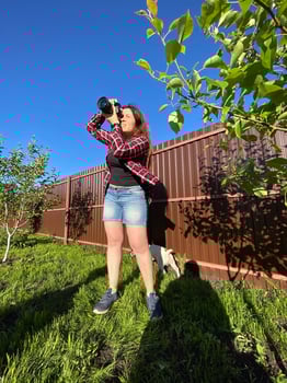 Millennial woman taking photo in a sunny backyard with a small tree and pet dog. Bright and clear blue sky providing a perfect backdrop for outdoor photography. Photographer, connection with nature
