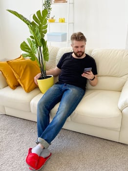 Bearded man relaxing on a cozy white couch with a smartphone and artificial interior plant. Modern living apartment with casual comfort and minimalism style. photo