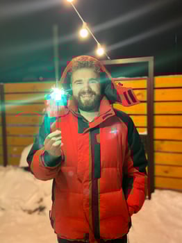 Bearded man in a red jacket holds a glowing sparkler, standing outside on a snowy winter night, with festive string lights hanging overhead. Christmas and New Year celebration concept photo