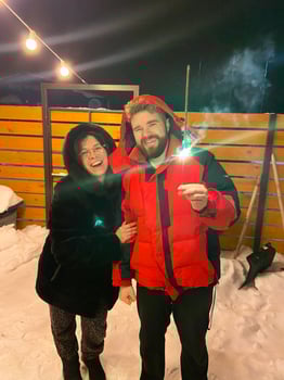 Bearded man in a red jacket with woman in fur coat holds a glowing sparkler, standing outside on a snowy winter night, with festive string lights hanging overhead. Christmas and New Year celebration photo