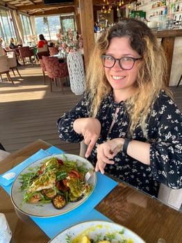 Woman enjoys a meal at a seaside restaurant, with a plate of grilled vegetables and chicken on toast. Bright and open atmosphere, surrounded by palm trees and a scenic view of beach outside photo