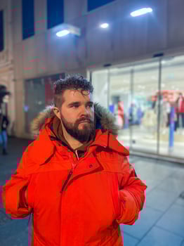 Man walking in a bustling nighttime city street, wearing a bright orange parka. Serene contemplation against the backdrop of lively city lights at night. Urban life concept photo