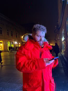 Millennial man in vivid red parka stands on city street at night, engrossed in their smartphone. Illuminated by the warm glow of city lights, capturing essence of urban life and digital connectivity. photo