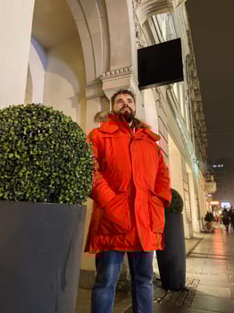 Millennial man in bright orange parka stands confidently on a city street, framed by architectural arches and urban greenery in cold season. Modern fashion and classic city charm photo