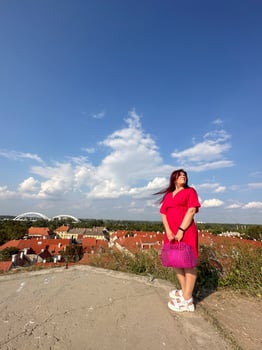 Woman in a pink dress standing on a hill overlooking a scenic town. Capturing the beauty of nature and architecture under a bright blue sky filled with fluffy clouds photo