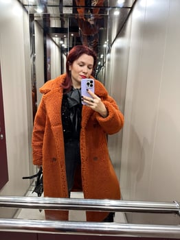 Fashionable woman wearing a vibrant orange winter coat takes a selfie in a modern elevator, capturing their stylish outfit and confident pose. Blog, influencer and blogger concept photo