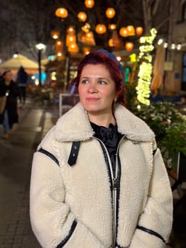 Millennial woman standing outdoors at Christmas night, wrapped in a cozy shearling jacket. the city lights in the background create a warm and Christmas holidays atmosphere, enhancing photo