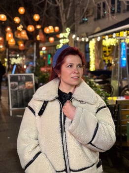 Millennial woman standing outdoors at Christmas night, wrapped in a cozy shearling jacket. the city lights in the background create a warm and Christmas holidays atmosphere, enhancing photo