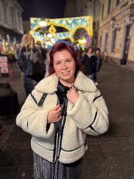 Millennial woman standing outdoors at Christmas night, wrapped in a cozy shearling jacket. the city lights in the background create a warm and Christmas holidays atmosphere, enhancing the peaceful photo