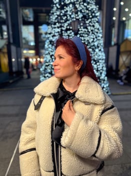 Millennial woman standing outdoors at Christmas night, wrapped in a cozy shearling jacket. the city lights in the background create a warm and Christmas holidays atmosphere, enhancing photo
