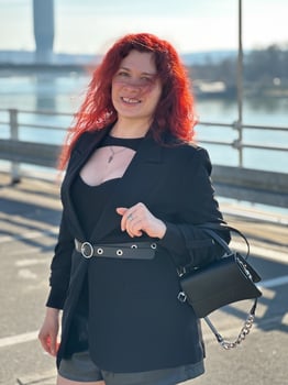 Millennial woman with long curly red hair gazing thoughtfully at the camera on a sunny day. Background features modern buildings and a river, evoking a sense of urban calm photo