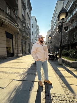 Millennial man stands confidently on a sunlit city street, wearing a cozy jacket and stylish hat. Urban landscape and soft morning light create a scene of casual elegance and modern lifestyle photo