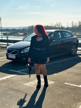 Millennial woman with long curly red hair gazing thoughtfully at the camera on a sunny day. Background features black car and a river, evoking a sense of urban calm. photo