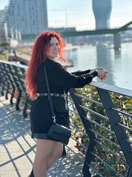 Millennial woman with long curly red hair leans on a black railing, gazing thoughtfully at the camera on a sunny day. Background features modern buildings and a river, evoking a sense of urban calm photo