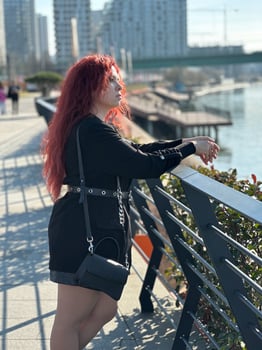 Millennial woman with long curly red hair leans on a black railing, gazing thoughtfully at the camera on a sunny day. Background features modern buildings and a river, evoking a sense of urban calm photo