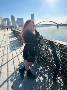 Millennial woman with long curly red hair leans on a black railing, gazing thoughtfully at the camera on a sunny day. Background features modern buildings and a river, evoking a sense of urban calm photo