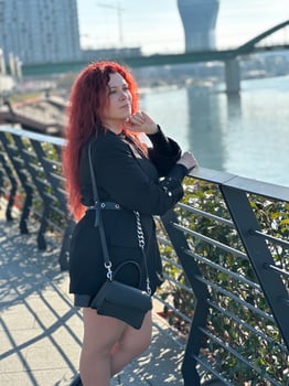 Millennial woman with long curly red hair leans on a black railing, gazing thoughtfully at the camera on a sunny day. Background features modern buildings and a river, evoking a sense of urban calm photo