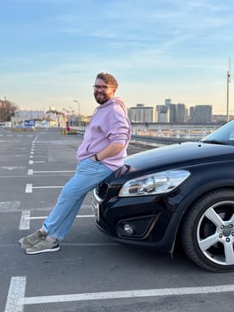 Millennial man in a casual hoodie leans confidently against a sleek black car in a parking lot. Peaceful evening sky forms a serene backdrop to this moment of relaxation and contemplation photo