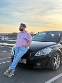 Millennial man in a casual hoodie leans confidently against a sleek black car in a parking lot. Peaceful evening sky forms a serene backdrop to this moment of relaxation and contemplation photo