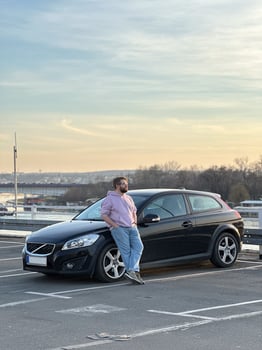 Millennial man in a casual hoodie leans confidently against a sleek black car in a parking lot. Peaceful evening sky forms a serene backdrop to this moment of relaxation and contemplation photo