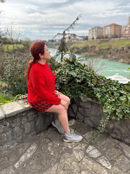 Millennial woman in a red sweater sitting on a stone ledge, enjoying a tranquil river view with a backdrop of city buildings. Autumn scene photo