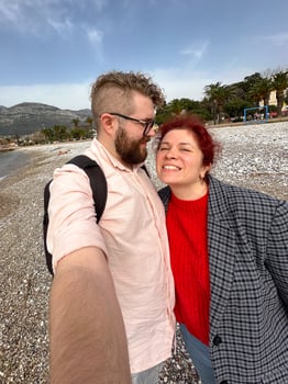 couple takes a cheerful selfie on a pebbled beach, with vast mountains and a serene sea in the background. their relaxed expressions and casual attire complement the picturesque coastal scenery photo