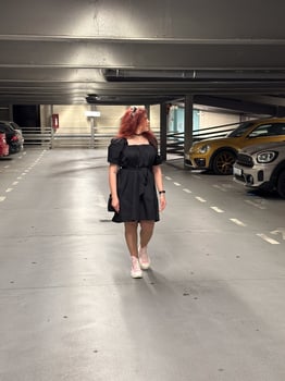 Millennial woman in a black dress walks through an empty parking garage, her pink sneakers adding a pop of color. Setting is urban and modern, with cars lined up neatly in the dimly lit space photo