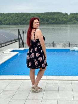 Woman with red hair in a black dress stands by a poolside, smiling and looking back at the camera. Happy vacation moment with a lush green river bank in background photo