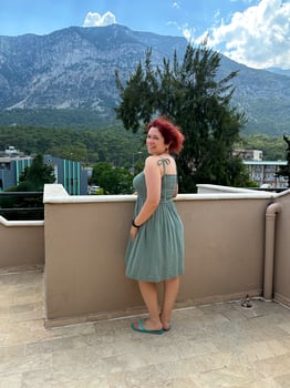 Millennial woman in a green dress stands on a rooftop terrace, gazing at the majestic mountains under a sky filled with fluffy clouds. Vacations and travel concept photo