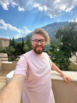 Millennial man with glasses and curly hair is taking a selfie from a scenic terrace. Beautiful mountain landscape under a partly cloudy sky creates a background. photo