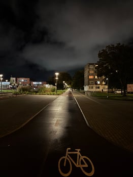 The image shows a night scene of an urban area with a paved road leading towards buildings in the distance. There is a bicycle lane symbol painted on the road, and street lights illuminate the scene. photo