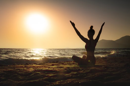 Back view of young Caucasian woman in lotus position sitting on sandy beach by the sea at sunrise or sunset. Sun salutation yoga practice photo
