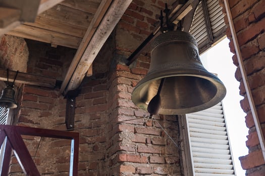 Large metal bell hanging inside a structure with brick walls and wooden beams. The architecture has a rustic feel, reminiscent of traditional or historical buildings. photo