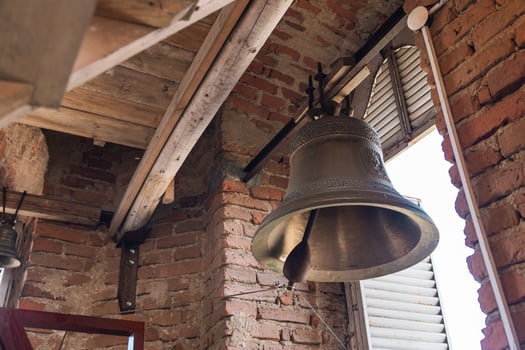 Large metal bell hanging inside a structure with brick walls and wooden beams. The architecture has a rustic feel, reminiscent of traditional or historical buildings. photo